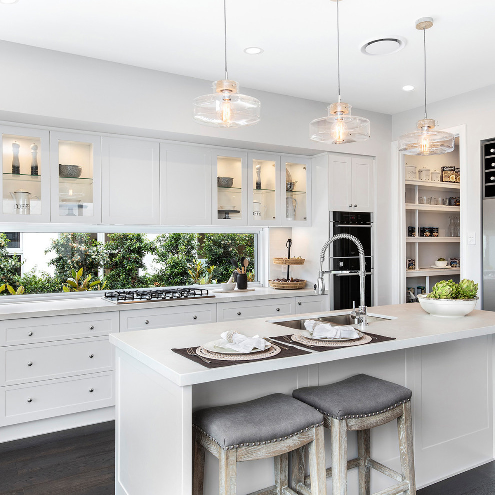 This is an example of a large transitional galley kitchen in Sydney with a double-bowl sink, shaker cabinets, white cabinets, quartz benchtops, black appliances, dark hardwood floors, with island, brown floor, white benchtop and window splashback.