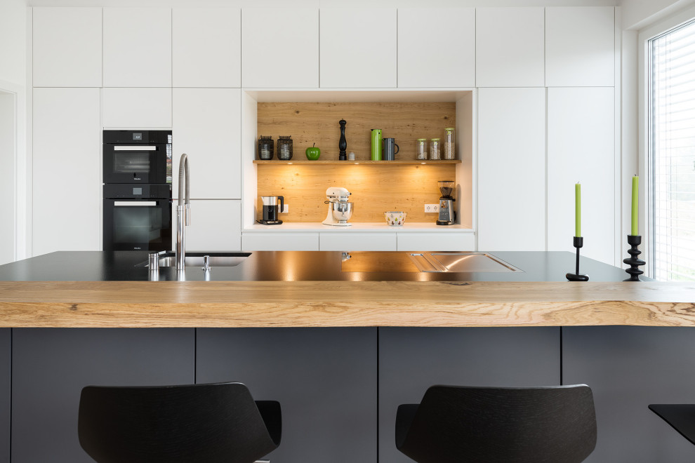 This is an example of a large contemporary eat-in kitchen in Munich with an undermount sink, flat-panel cabinets, white cabinets, granite benchtops, brown splashback, timber splashback, black appliances, medium hardwood floors, with island, brown floor and black benchtop.