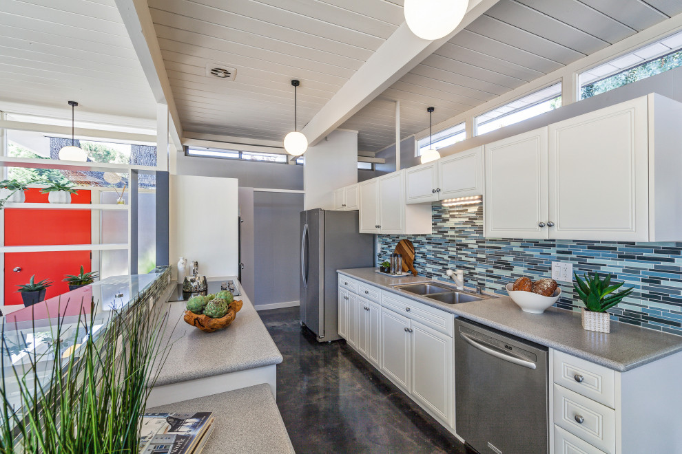 Midcentury galley kitchen in San Francisco with an undermount sink, raised-panel cabinets, white cabinets, blue splashback, mosaic tile splashback, stainless steel appliances, concrete floors, with island, black floor, grey benchtop, exposed beam and timber.