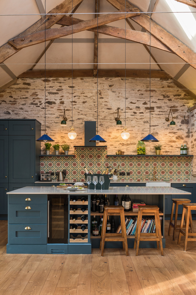 Transitional kitchen in London with cement tile splashback, a farmhouse sink, shaker cabinets, blue cabinets, multi-coloured splashback, light hardwood floors, with island and grey benchtop.