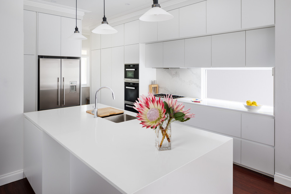 Contemporary kitchen in Perth with an undermount sink, flat-panel cabinets, white cabinets, white splashback, stainless steel appliances, with island, brown floor and white benchtop.