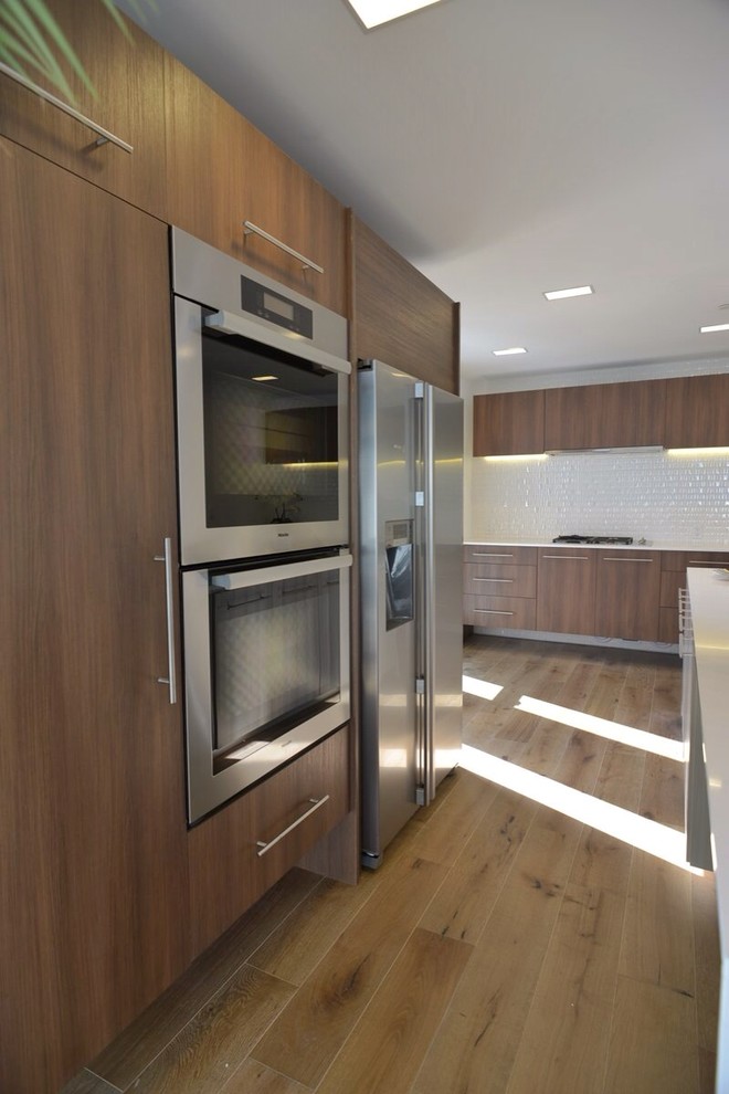 Design ideas for a large modern l-shaped open plan kitchen in Los Angeles with a drop-in sink, flat-panel cabinets, medium wood cabinets, solid surface benchtops, white splashback, porcelain splashback, stainless steel appliances, light hardwood floors and with island.