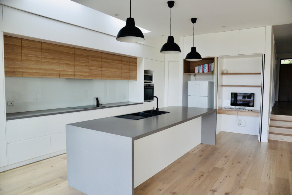 Inspiration for a large scandi galley open plan kitchen in Melbourne with a double-bowl sink, flat-panel cabinets, white cabinets, composite countertops, white splashback, glass sheet splashback, stainless steel appliances, light hardwood flooring, an island, beige floors, grey worktops and a drop ceiling.