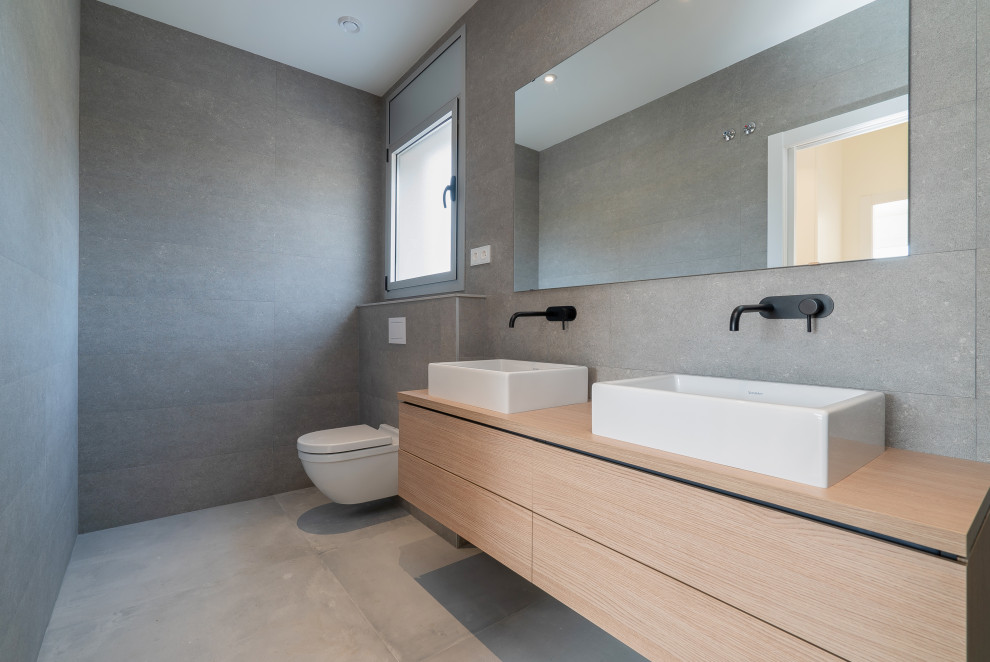 This is an example of a mid-sized contemporary 3/4 bathroom in Barcelona with recessed-panel cabinets, brown cabinets, an alcove tub, a shower/bathtub combo, a wall-mount toilet, gray tile, ceramic tile, grey walls, ceramic floors, a vessel sink, wood benchtops, grey floor, an open shower, brown benchtops, an enclosed toilet, a single vanity, a built-in vanity, wallpaper and panelled walls.