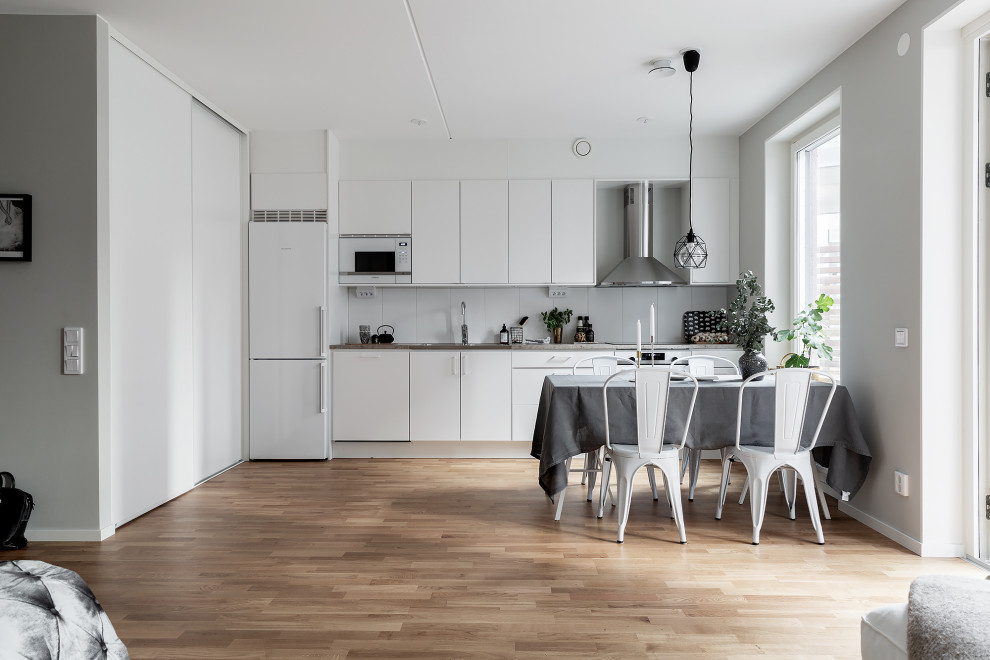 This is an example of a mid-sized scandinavian single-wall eat-in kitchen in Stockholm with a triple-bowl sink, flat-panel cabinets, white cabinets, white splashback, porcelain splashback, stainless steel appliances, medium hardwood floors, no island, brown floor and grey benchtop.