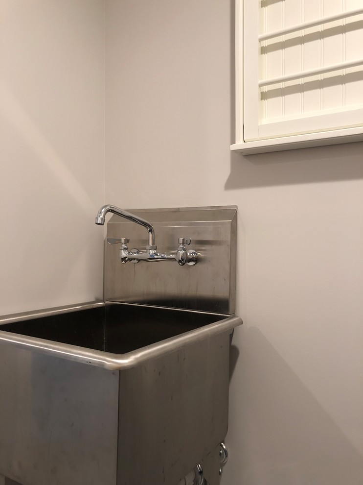 Inspiration for a large transitional l-shaped dedicated laundry room in New York with a farmhouse sink, shaker cabinets, white cabinets, quartz benchtops, white splashback, ceramic splashback, dark hardwood floors, brown floor, white benchtop, grey walls and a side-by-side washer and dryer.