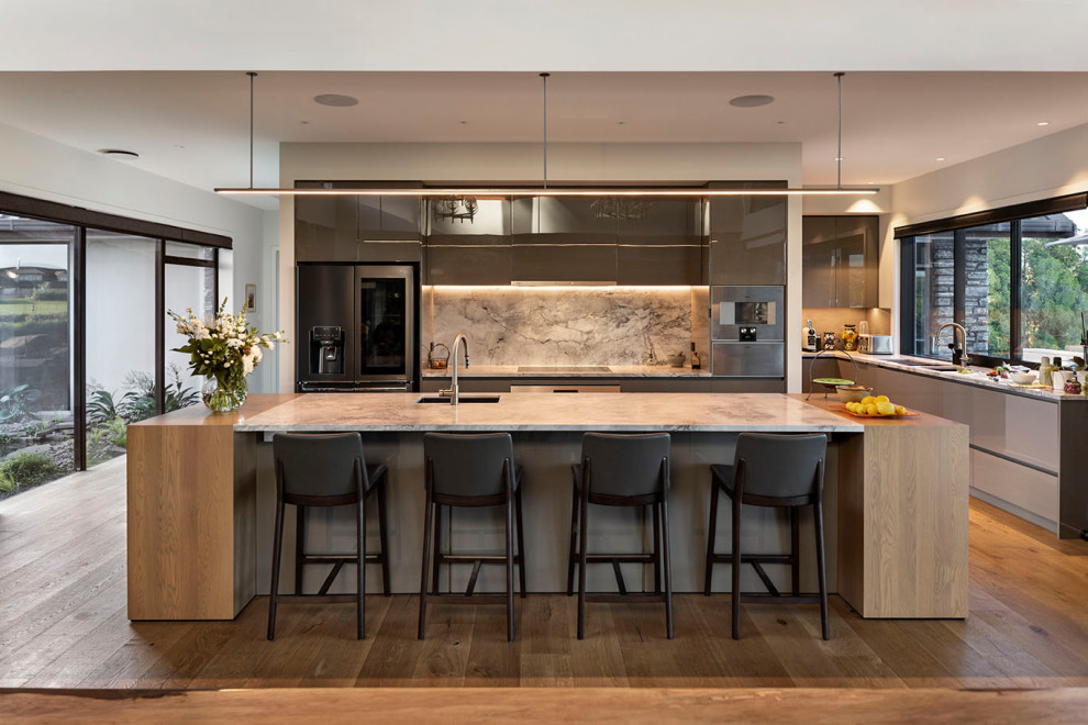 Design ideas for a large contemporary l-shaped open plan kitchen in Auckland with an undermount sink, recessed-panel cabinets, grey cabinets, granite benchtops, grey splashback, granite splashback, stainless steel appliances, medium hardwood floors, with island, brown floor and white benchtop.