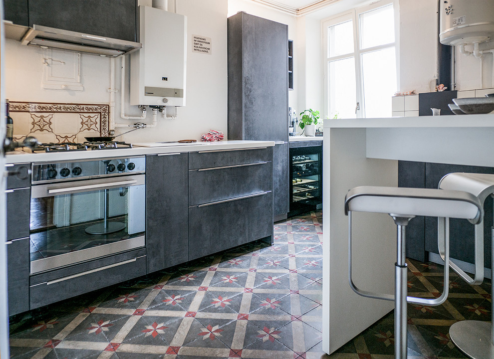 Photo of a mid-sized industrial galley separate kitchen in Other with a single-bowl sink, grey cabinets, solid surface benchtops, white splashback, cement tile splashback, stainless steel appliances, cement tiles, no island, multi-coloured floor and white benchtop.
