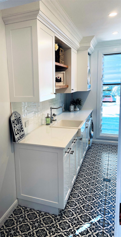 Photo of a large beach style galley dedicated laundry room in Brisbane with a farmhouse sink, shaker cabinets, white cabinets, quartz benchtops, white splashback, subway tile splashback, white walls, ceramic floors, black floor, white benchtop, recessed and panelled walls.