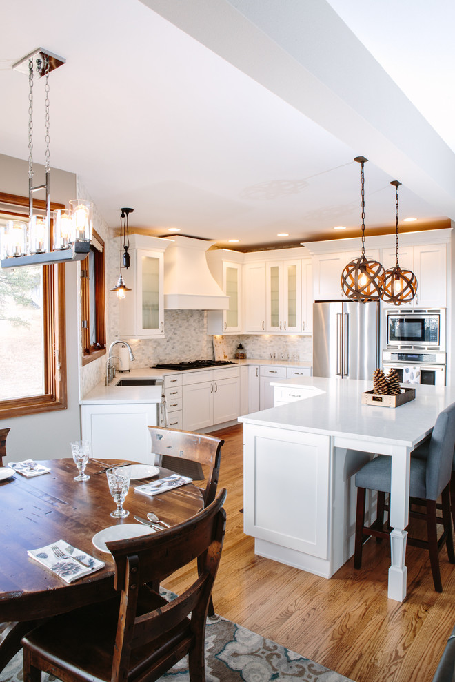 This is an example of a mid-sized transitional u-shaped eat-in kitchen in Denver with a farmhouse sink, shaker cabinets, white cabinets, quartz benchtops, white splashback, stone tile splashback, stainless steel appliances, medium hardwood floors, with island and brown floor.