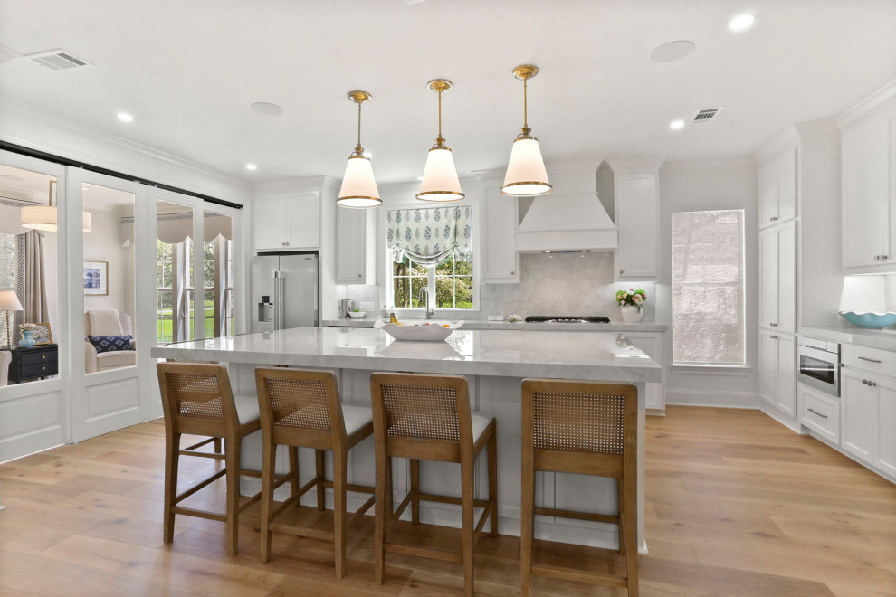 Example of a large transitional l-shaped light wood floor and brown floor open concept kitchen design in New Orleans with an undermount sink, shaker cabinets, white cabinets, quartz countertops, white backsplash, quartz backsplash, stainless steel appliances, an island and white countertops