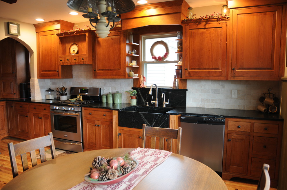Photo of a mid-sized country l-shaped eat-in kitchen in Columbus with a farmhouse sink, raised-panel cabinets, medium wood cabinets, beige splashback, stone tile splashback, stainless steel appliances, light hardwood floors, no island and soapstone benchtops.