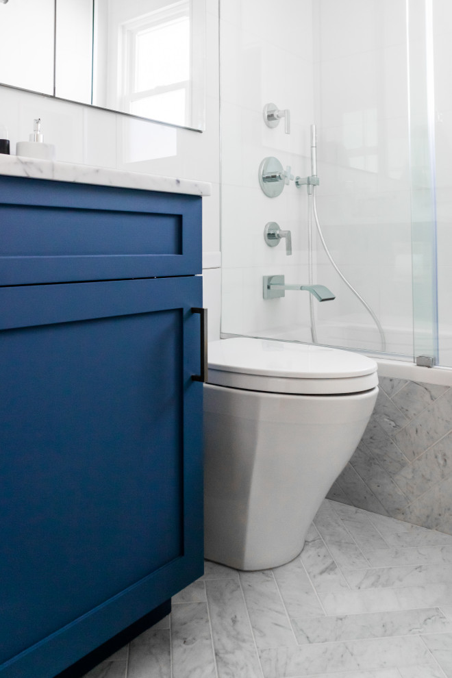 Inspiration for a small transitional bathroom in New York with shaker cabinets, blue cabinets, a drop-in tub, a shower/bathtub combo, a one-piece toilet, white tile, porcelain tile, white walls, marble floors, an undermount sink, marble benchtops, white floor, a sliding shower screen and white benchtops.