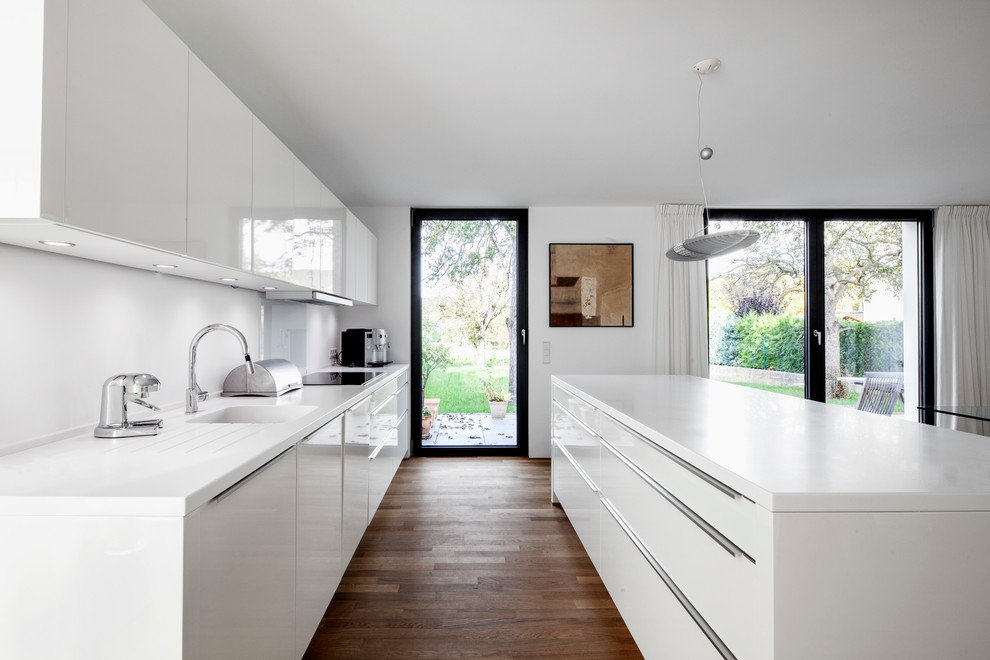 Mid-sized contemporary single-wall open plan kitchen in Cologne with with island, an integrated sink, flat-panel cabinets, white cabinets, white splashback, glass sheet splashback, panelled appliances and dark hardwood floors.