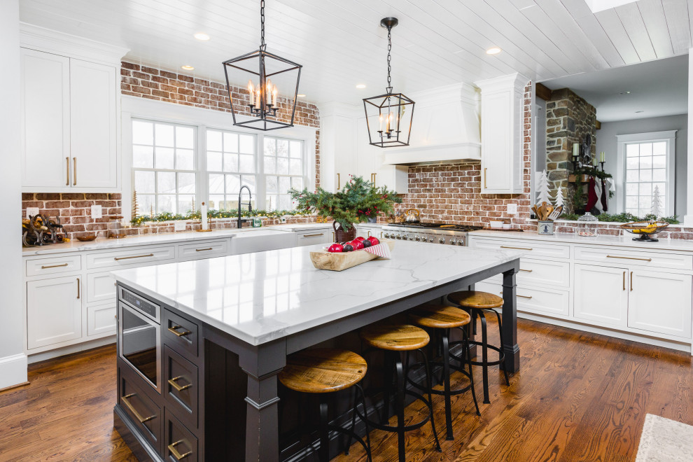 This is an example of a large traditional kitchen in DC Metro with a farmhouse sink, shaker cabinets, white cabinets, quartz benchtops, red splashback, brick splashback, stainless steel appliances, with island, white benchtop and medium hardwood floors.