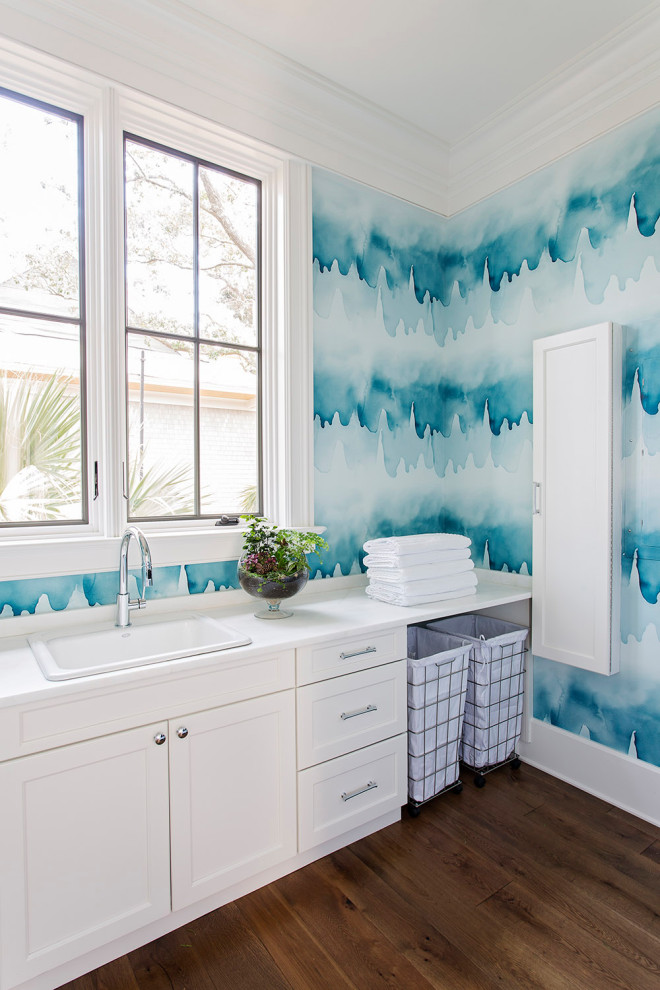 Beach style single-wall dedicated laundry room in Charleston with a drop-in sink, shaker cabinets, white cabinets, blue walls, dark hardwood floors, brown floor, white benchtop and wallpaper.
