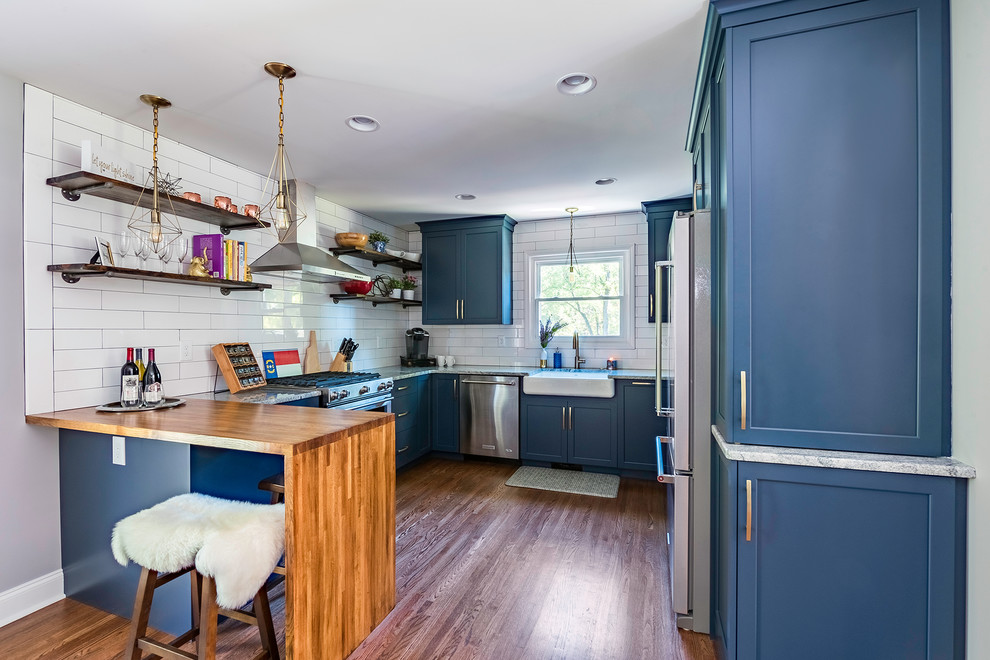This is an example of a mid-sized eclectic u-shaped eat-in kitchen in Charlotte with a farmhouse sink, shaker cabinets, blue cabinets, granite benchtops, white splashback, ceramic splashback, stainless steel appliances, medium hardwood floors, a peninsula and brown floor.