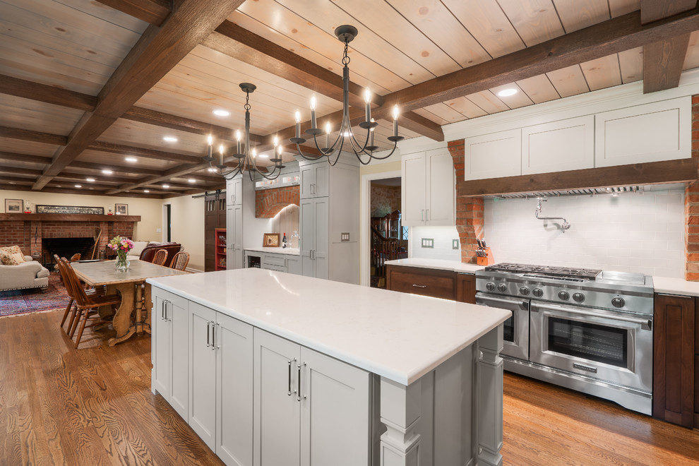 Example of a mid-sized transitional galley medium tone wood floor and brown floor eat-in kitchen design in Columbus with a farmhouse sink, shaker cabinets, quartz countertops, white backsplash, subway tile backsplash, stainless steel appliances, an island, white countertops and gray cabinets