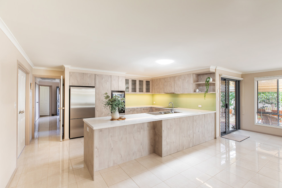 This is an example of a mid-sized modern u-shaped kitchen pantry in Adelaide with a double-bowl sink, flat-panel cabinets, light wood cabinets, quartz benchtops, green splashback, glass sheet splashback, stainless steel appliances, porcelain floors, a peninsula, white floor and white benchtop.