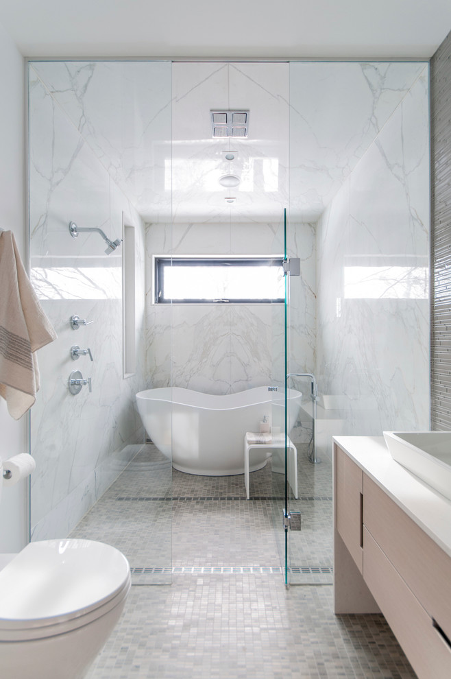 This is an example of a mid-sized contemporary master wet room bathroom in Toronto with flat-panel cabinets, light wood cabinets, a freestanding tub, a one-piece toilet, white tile, stone slab, white walls, mosaic tile floors, a vessel sink and a hinged shower door.
