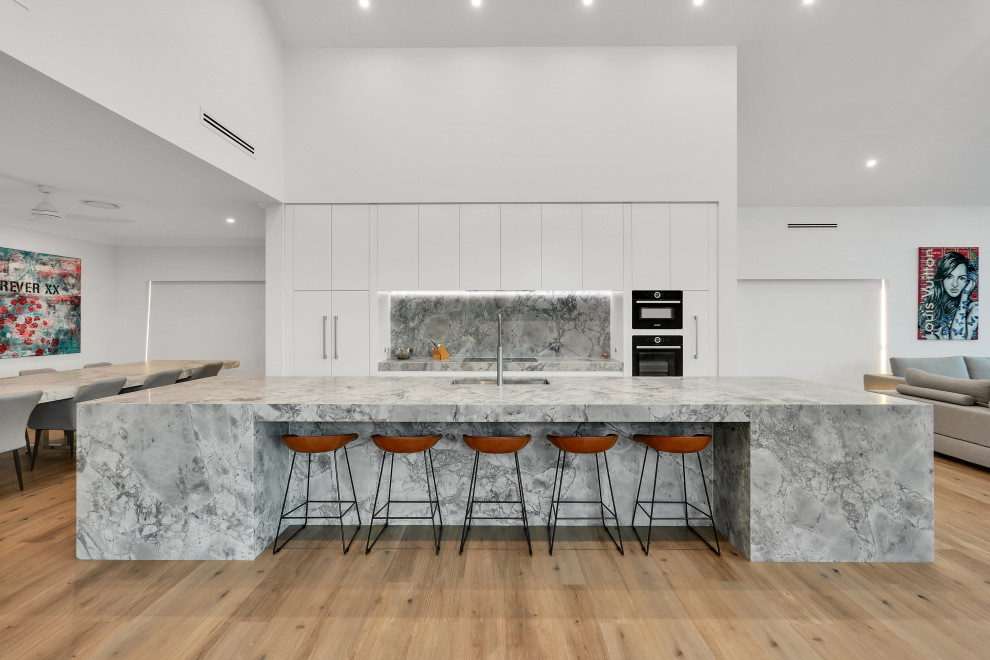 Design ideas for an expansive modern grey and white galley open plan kitchen in Sydney with a submerged sink, flat-panel cabinets, white cabinets, black appliances, an island, beige floors, grey worktops, a vaulted ceiling, granite worktops, grey splashback, granite splashback and vinyl flooring.
