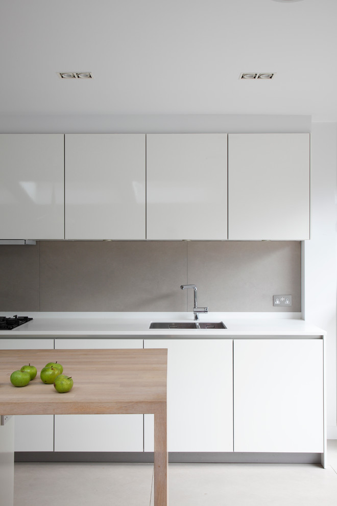 Photo of a mid-sized contemporary single-wall open plan kitchen in London with flat-panel cabinets, with island, an integrated sink, white cabinets, solid surface benchtops, grey splashback, ceramic splashback, panelled appliances, ceramic floors, grey floor, white benchtop and vaulted.