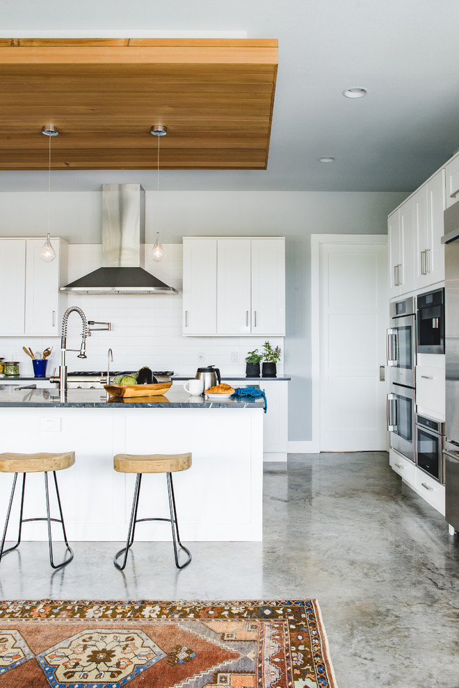 Photo of a contemporary galley kitchen in Austin with a farmhouse sink, flat-panel cabinets, white cabinets, white splashback, subway tile splashback, stainless steel appliances, concrete floors and with island.
