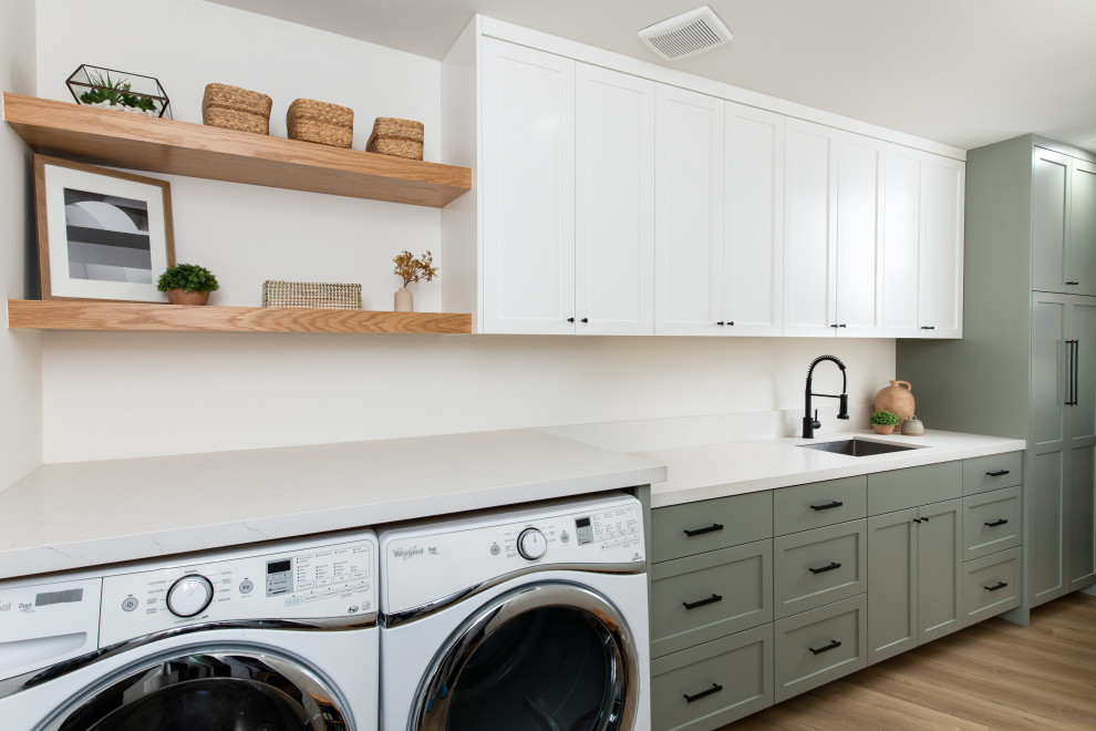 This is an example of a medium sized nautical separated utility room in Orange County with a built-in sink, green cabinets, engineered stone countertops, white splashback, engineered quartz splashback, white walls, medium hardwood flooring, a side by side washer and dryer and white worktops.