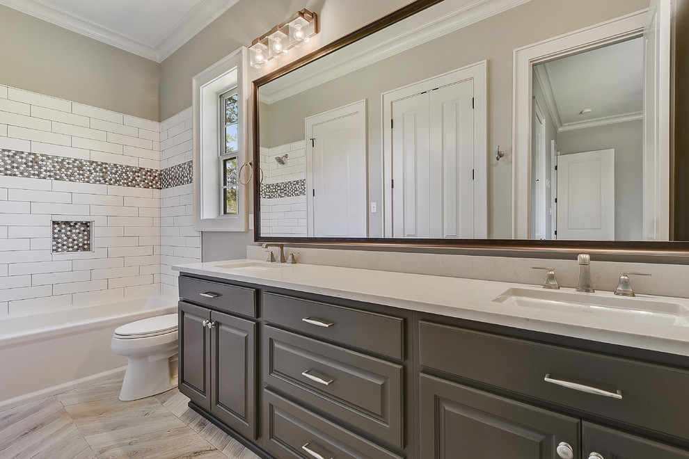 This is an example of a mid-sized traditional 3/4 bathroom in New Orleans with subway tile, engineered quartz benchtops, raised-panel cabinets, grey cabinets, an alcove tub, a shower/bathtub combo, white tile, beige walls, light hardwood floors, an undermount sink, brown floor and white benchtops.