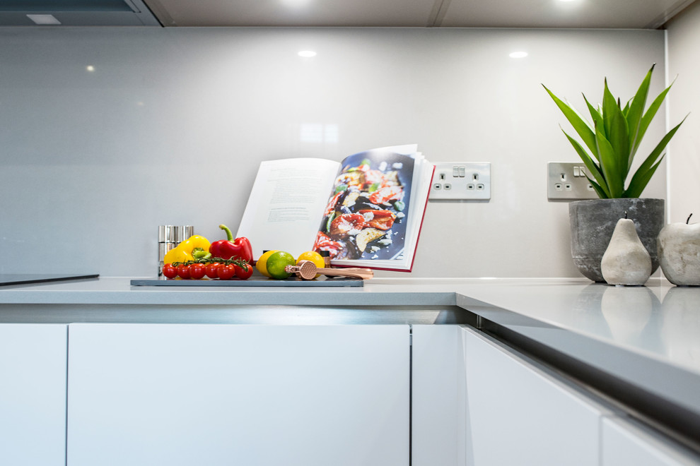 This is an example of a contemporary eat-in kitchen in Other with a single-bowl sink, flat-panel cabinets, white cabinets, quartzite benchtops, grey splashback, glass sheet splashback, black appliances, porcelain floors, with island, beige floor and white benchtop.