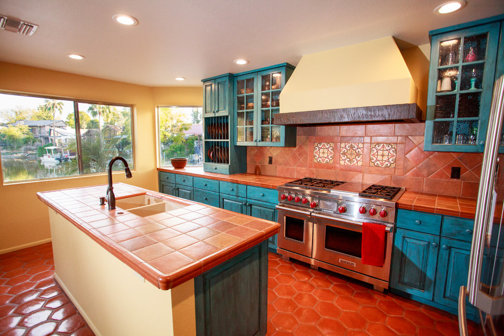 Photo of a country l-shaped kitchen in Phoenix with an undermount sink, raised-panel cabinets, blue cabinets, tile benchtops, multi-coloured splashback, ceramic splashback, stainless steel appliances, terra-cotta floors and with island.