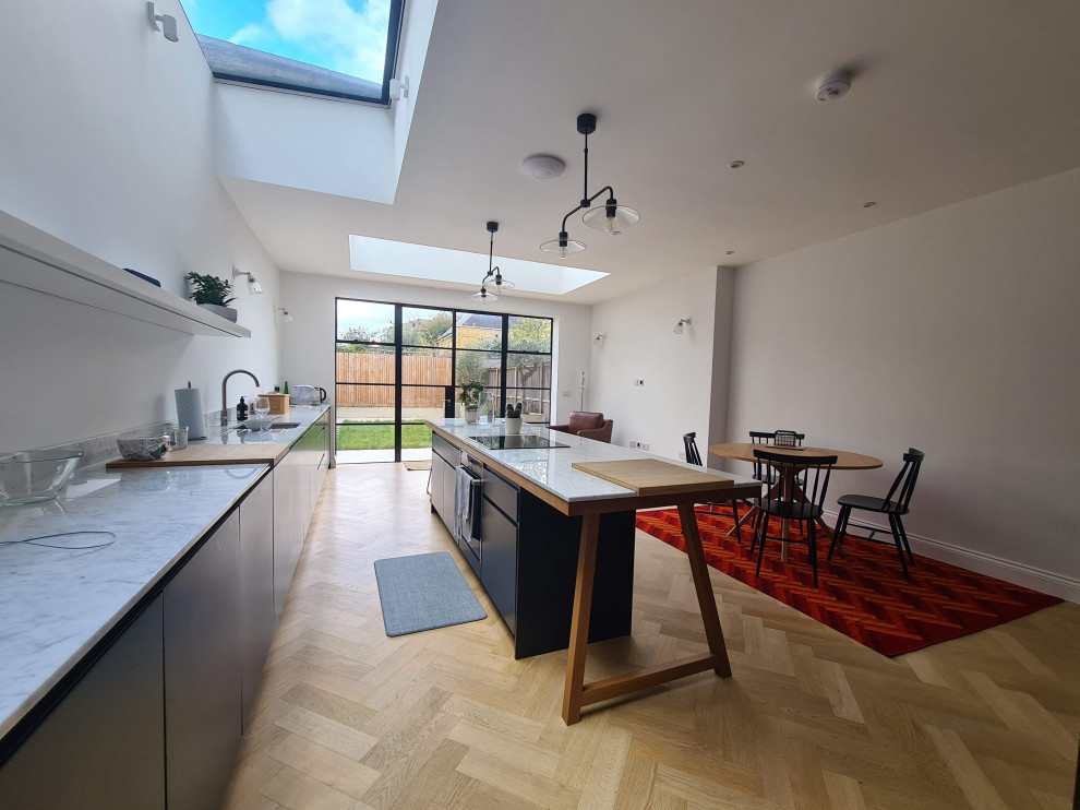 Inspiration for an expansive traditional l-shaped open plan kitchen in London with a drop-in sink, flat-panel cabinets, black cabinets, granite benchtops, white splashback, glass sheet splashback, black appliances, light hardwood floors, with island, brown floor, white benchtop and recessed.