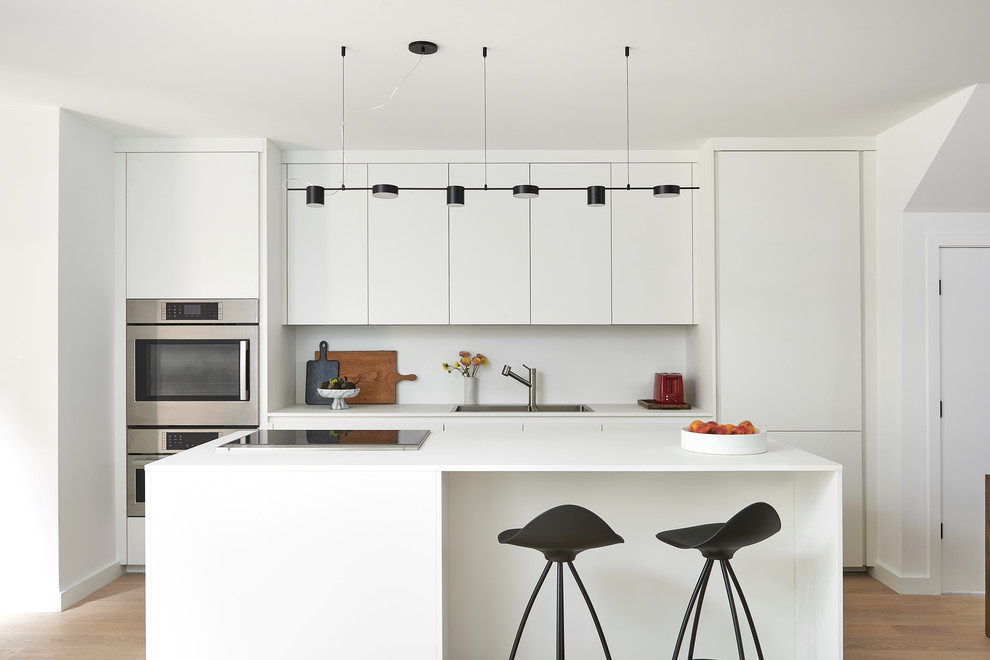 This is an example of a contemporary galley kitchen in Toronto with a drop-in sink, flat-panel cabinets, white cabinets, white splashback, stainless steel appliances, light hardwood floors, with island, beige floor and white benchtop.