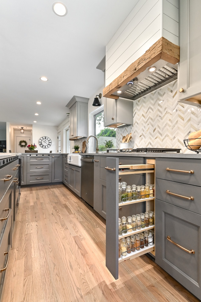 Inspiration for an expansive country l-shaped kitchen/diner in Milwaukee with a belfast sink, shaker cabinets, grey cabinets, engineered stone countertops, white splashback, marble splashback, stainless steel appliances, medium hardwood flooring, an island, brown floors and grey worktops.