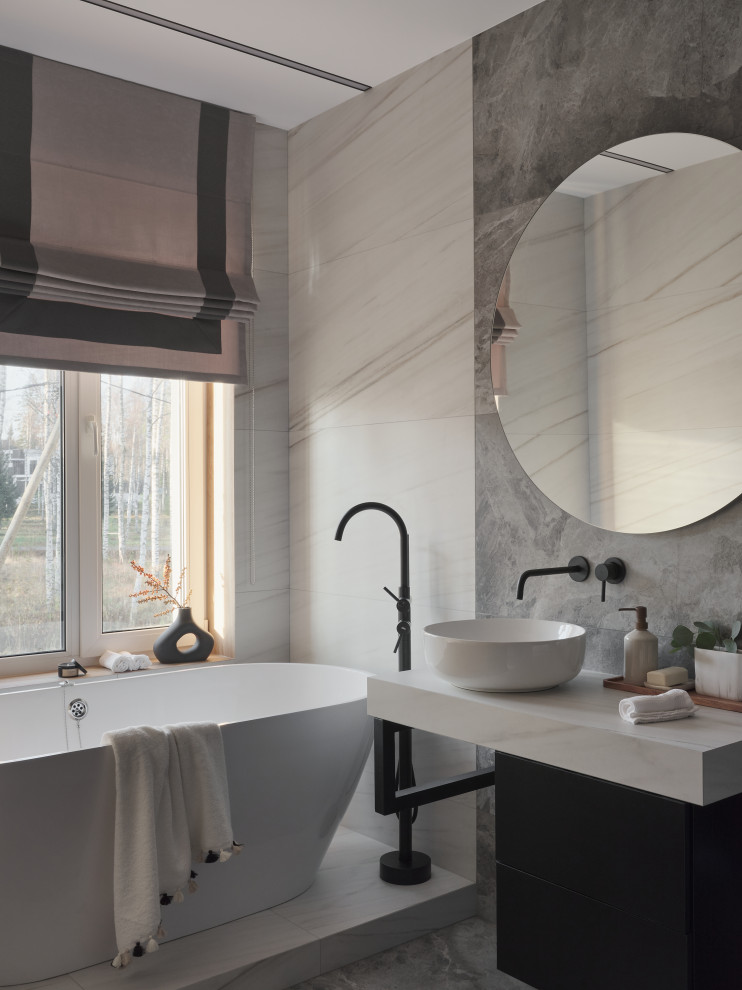 Идея дизайна: главная, серо-белая ванная комната среднего размера в современном стиле с плоскими фасадами, черными фасадами, отдельно стоящей ванной, инсталляцией, серой плиткой, керамогранитной плиткой, серыми стенами, полом из керамогранита, накладной раковиной, серым полом, белой столешницей, окном, тумбой под одну раковину и подвесной тумбой