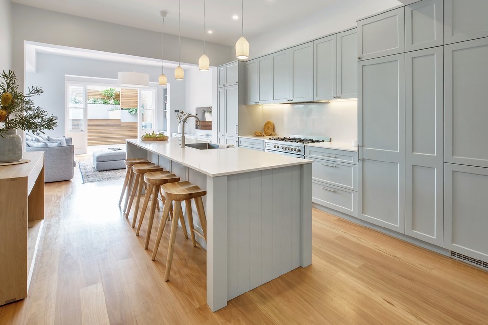 This is an example of a mid-sized beach style single-wall kitchen pantry in Sydney with a single-bowl sink, shaker cabinets, grey cabinets, solid surface benchtops, white splashback, stainless steel appliances, medium hardwood floors, with island, brown floor and white benchtop.