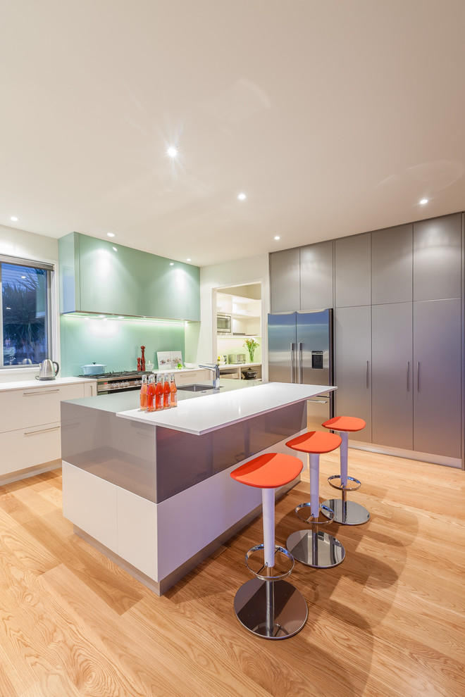 Design ideas for a mid-sized tropical l-shaped kitchen pantry in Auckland with an undermount sink, open cabinets, white cabinets, quartz benchtops, green splashback, glass sheet splashback, stainless steel appliances, light hardwood floors and with island.