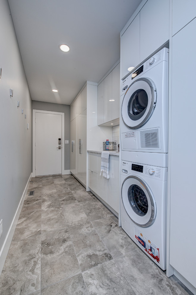 This is an example of a mid-sized contemporary single-wall dedicated laundry room in Calgary with an undermount sink, flat-panel cabinets, white cabinets, quartz benchtops, grey walls, limestone floors, a stacked washer and dryer, grey floor and white benchtop.
