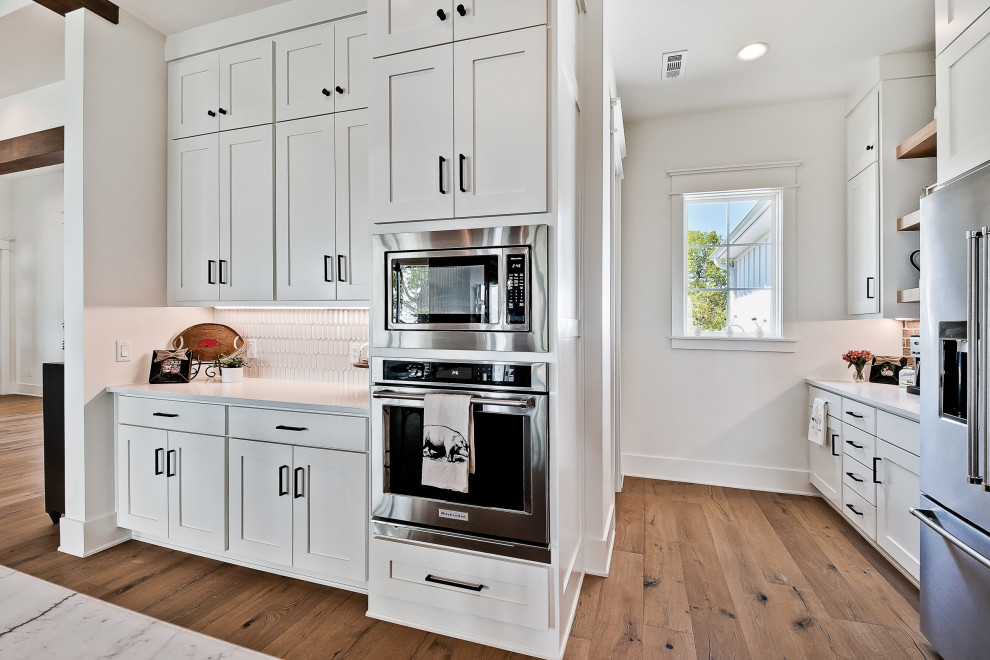 Design ideas for a large country u-shaped eat-in kitchen in Other with a farmhouse sink, raised-panel cabinets, white cabinets, quartz benchtops, red splashback, brick splashback, stainless steel appliances, light hardwood floors, with island, white benchtop and exposed beam.