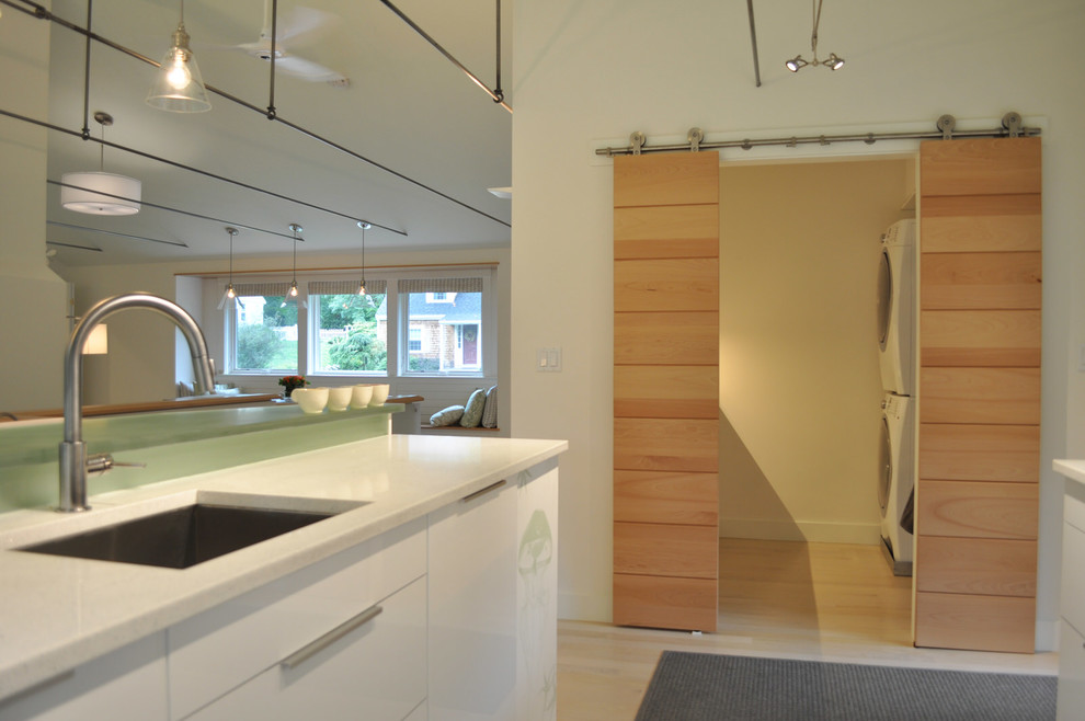 Photo of a small midcentury galley utility room in Boston with an undermount sink, flat-panel cabinets, white cabinets, solid surface benchtops, white walls, light hardwood floors and a stacked washer and dryer.