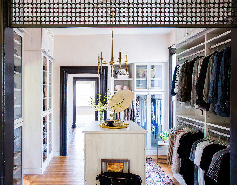 This is an example of an expansive transitional gender-neutral walk-in wardrobe in Other with glass-front cabinets, white cabinets and light hardwood floors.