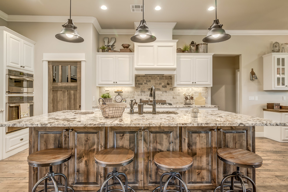 Design ideas for a mid-sized country l-shaped open plan kitchen in Oklahoma City with an undermount sink, raised-panel cabinets, white cabinets, granite benchtops, grey splashback, brick splashback, stainless steel appliances, medium hardwood floors and with island.