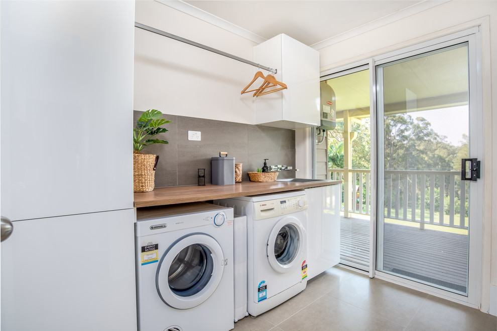 Design ideas for a mid-sized modern single-wall dedicated laundry room in Sunshine Coast with grey walls, ceramic floors, a drop-in sink, white cabinets, wood benchtops, a side-by-side washer and dryer, grey floor and brown benchtop.