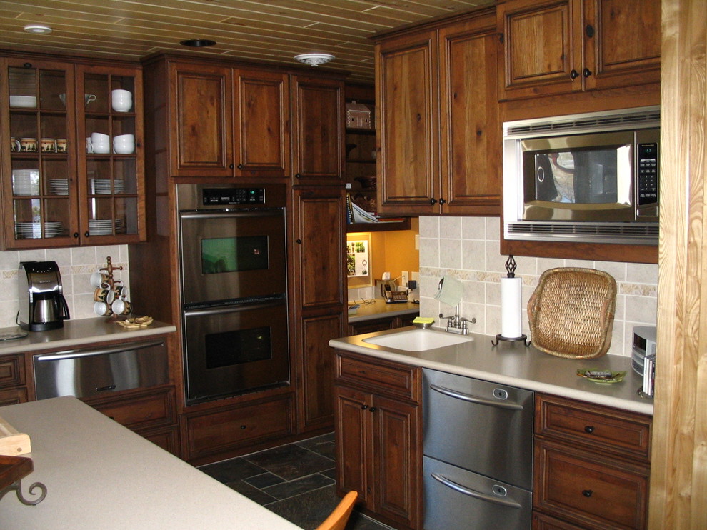 Enclosed kitchen - mid-sized rustic u-shaped slate floor and beige floor enclosed kitchen idea with an undermount sink, raised-panel cabinets, medium tone wood cabinets, quartz countertops, beige backsplash, stone tile backsplash, stainless steel appliances and an island