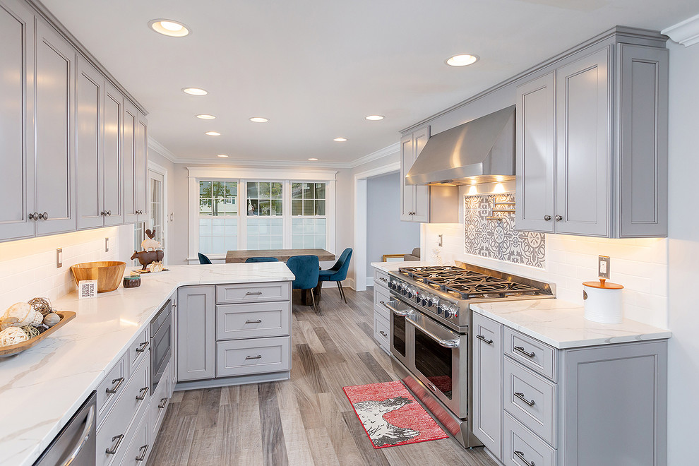This is an example of a large transitional u-shaped eat-in kitchen in Philadelphia with a farmhouse sink, raised-panel cabinets, grey cabinets, quartz benchtops, white splashback, ceramic splashback, stainless steel appliances, laminate floors, no island, brown floor and white benchtop.