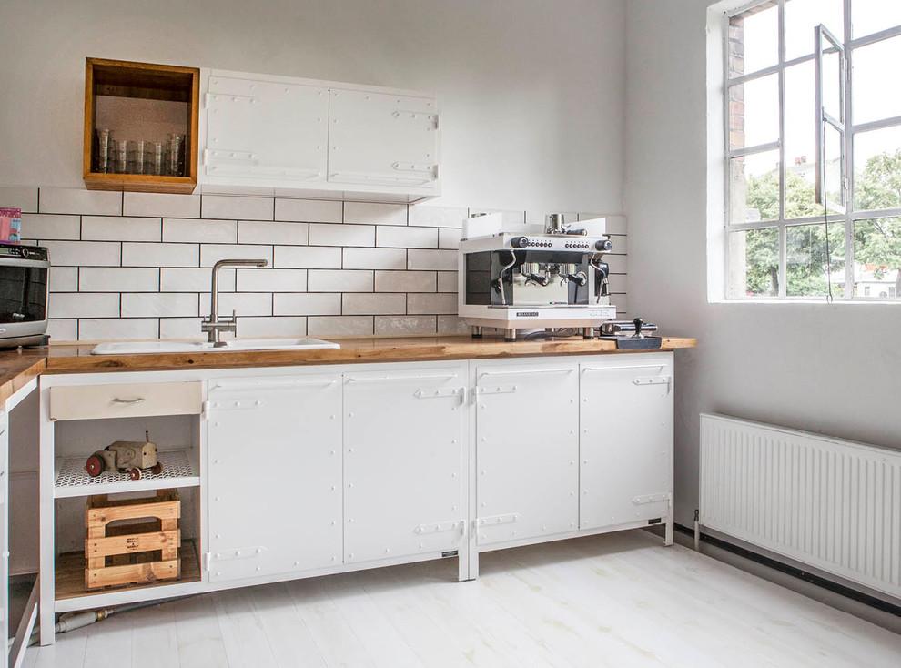 Design ideas for a mid-sized industrial l-shaped separate kitchen in Berlin with a drop-in sink, flat-panel cabinets, white cabinets, wood benchtops, white splashback, painted wood floors, no island, white floor, subway tile splashback and brown benchtop.