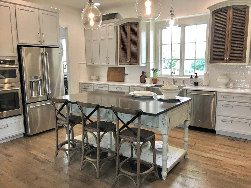 Example of a large classic u-shaped light wood floor and brown floor eat-in kitchen design with a farmhouse sink, flat-panel cabinets, gray cabinets, marble countertops, white backsplash, ceramic backsplash, stainless steel appliances and an island