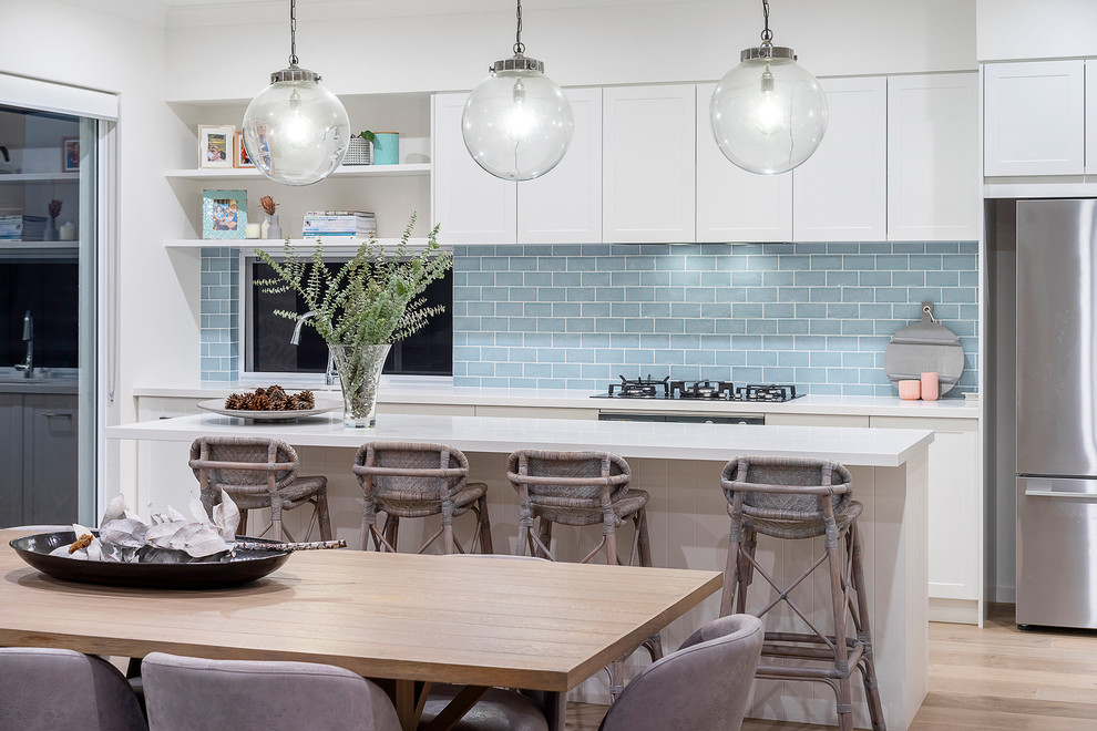 Photo of a mid-sized beach style galley eat-in kitchen in Perth with shaker cabinets, white cabinets, blue splashback, stainless steel appliances, light hardwood floors, with island, beige floor and white benchtop.