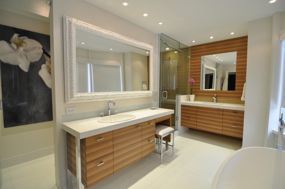 This is an example of a large contemporary master bathroom in Toronto with a drop-in sink, engineered quartz benchtops, a freestanding tub, an alcove shower, a one-piece toilet, glass tile, white walls, ceramic floors, flat-panel cabinets, medium wood cabinets, white floor, a hinged shower door and white benchtops.