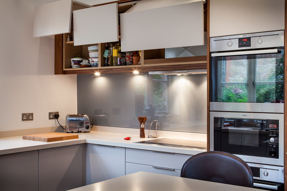 Design ideas for a mid-sized eclectic u-shaped eat-in kitchen in Hertfordshire with an undermount sink, flat-panel cabinets, grey cabinets, solid surface benchtops, grey splashback, glass sheet splashback, stainless steel appliances and a peninsula.